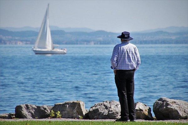 Älterer Mann mit Hut am See schaut Segelschiff nach